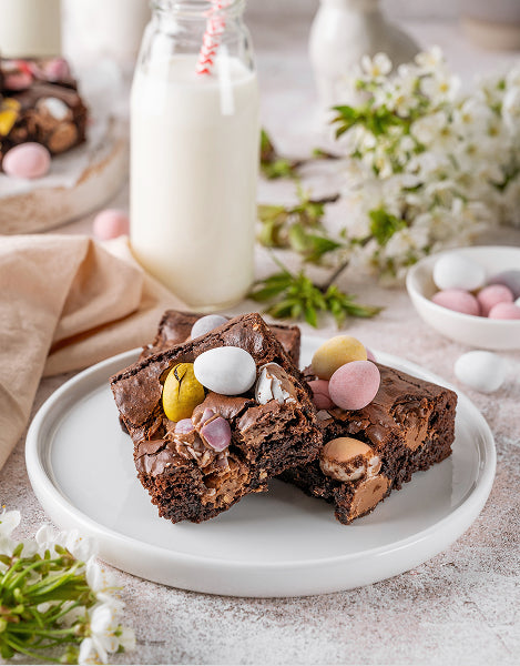 Brownies with Easter eggs on a plate with a glass of milk and flowers in the background.