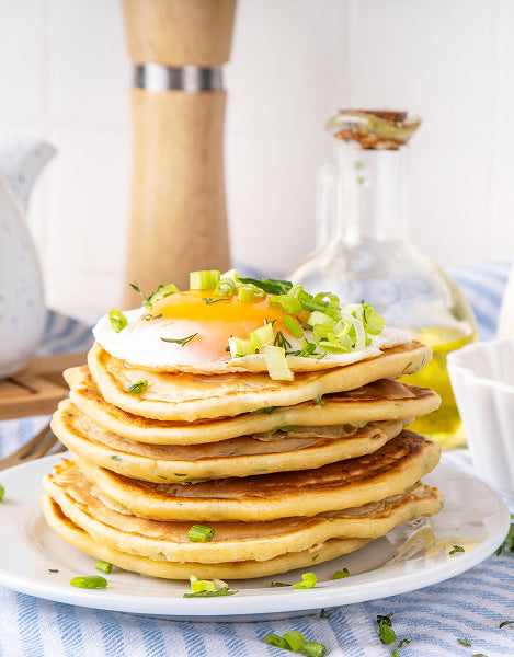 Stack of pancakes with a poached egg on a plate, garnished with green onions, against a blurred kitchen background.