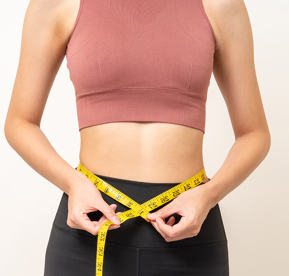 Woman measuring waist with a yellow tape measure against a plain background