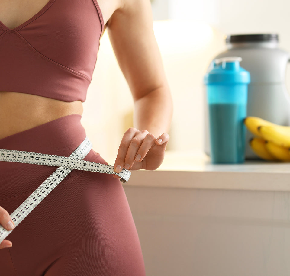 Woman measuring waist with a tape measure, with a shaker bottle and bananas in the background.