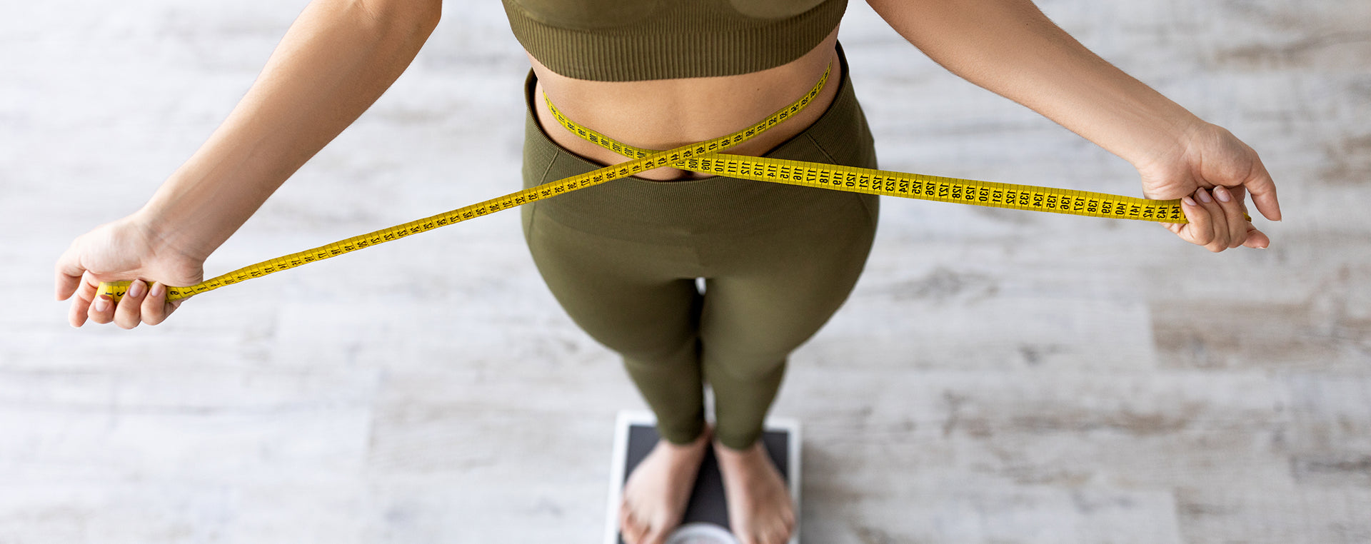 Person measuring waist with a tape measure on a scale against a light background